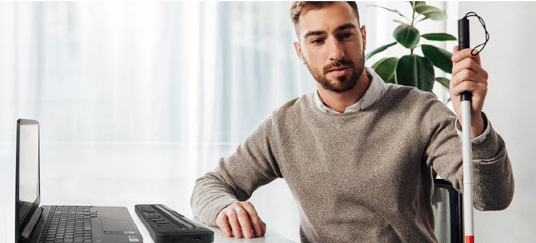 Un homme assis avec un ordinateur portable et son Brailliant BI 20X avec une canne blanche à côté de lui.