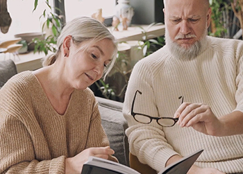 A man tries to read his wife's book by moving his glasses back and forth with his hand.