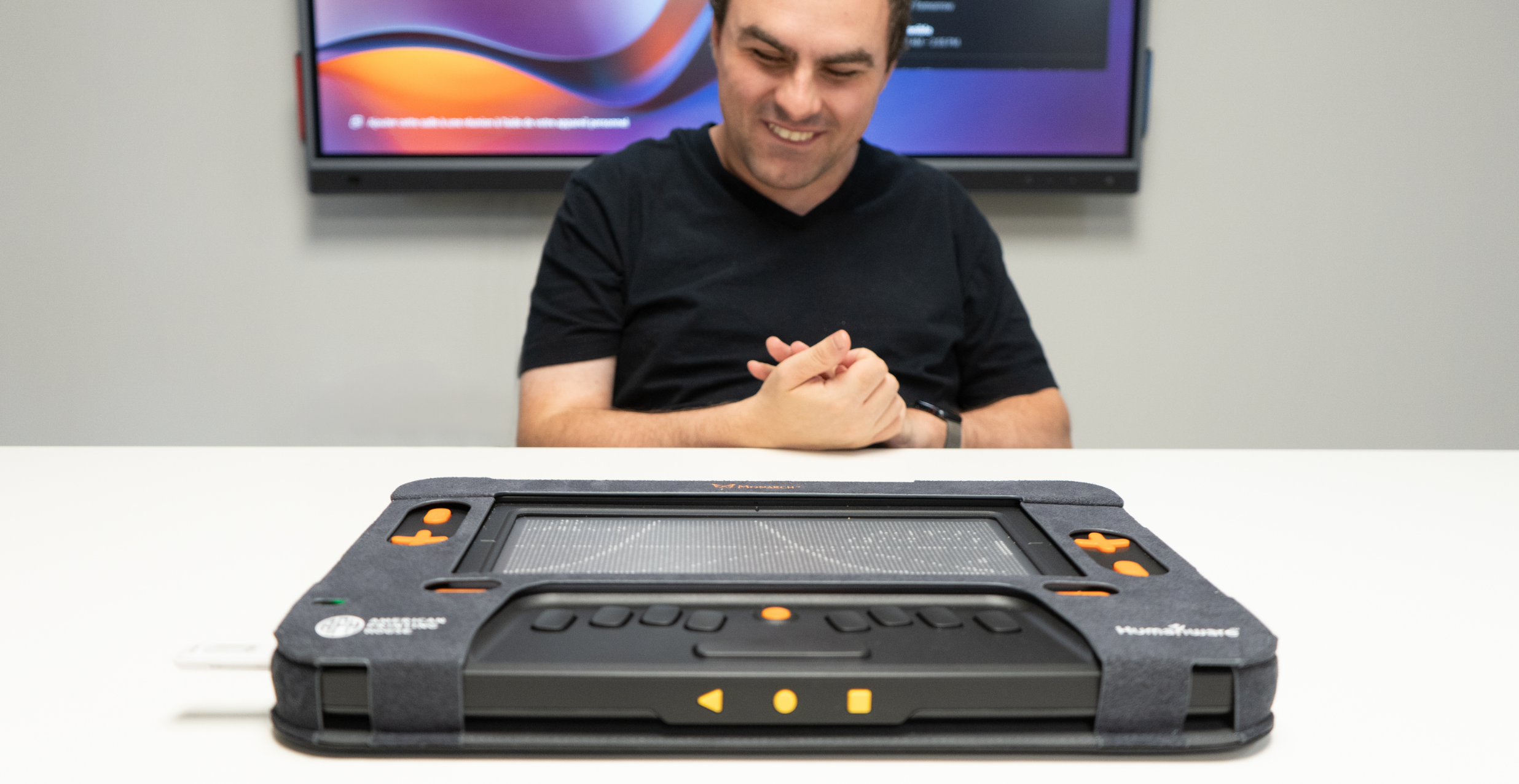 A man at a table in a conference room looks pleased as he uses both hands to touch the Monarch’s multiline braille display.