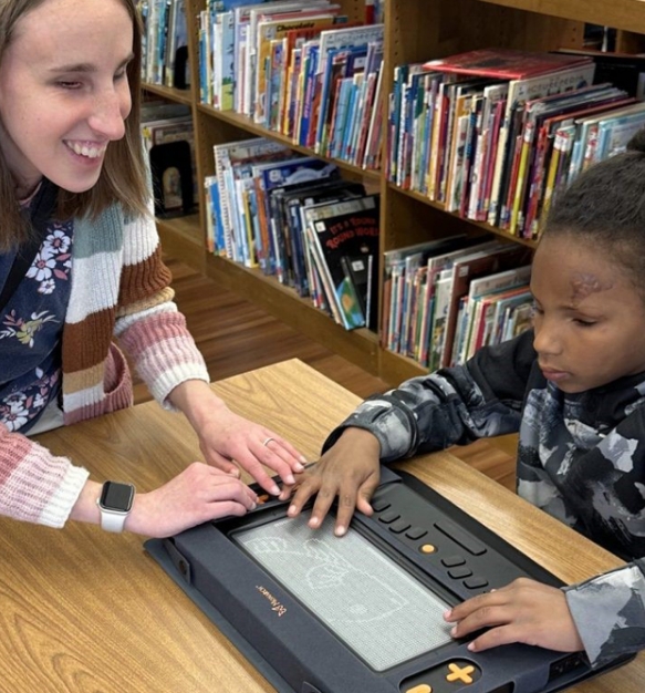 A woman in a striped sweater helps a young boy use the Monarch on a table in a school library.