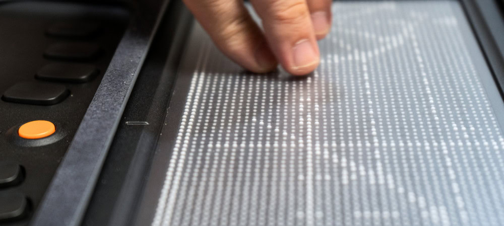 Image of a close-up shows a hand touching a tactile surface with Braille dots.