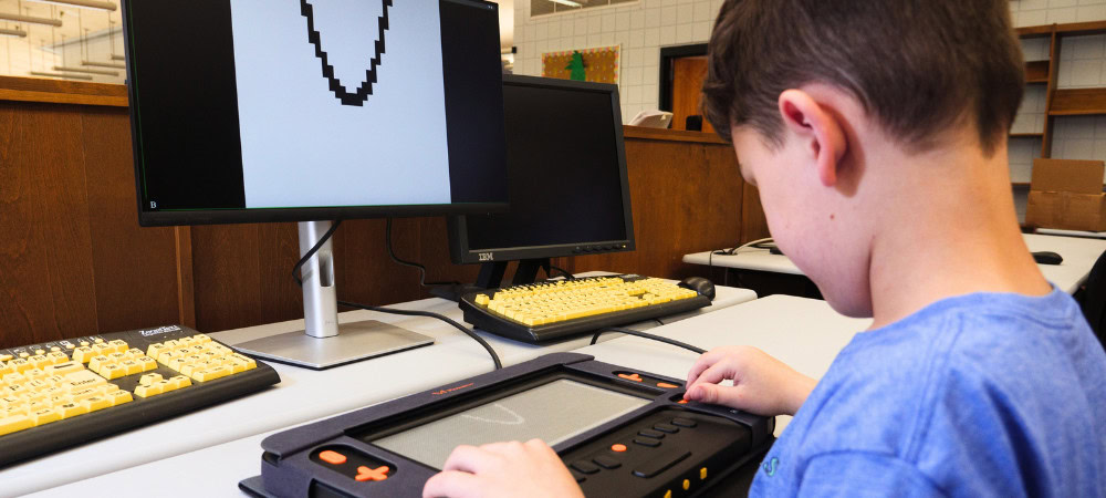 Image of a boy interacts with the Monarch in a library, a computer displays the simplistic image of a curve, highlighting tactile and visual learning.