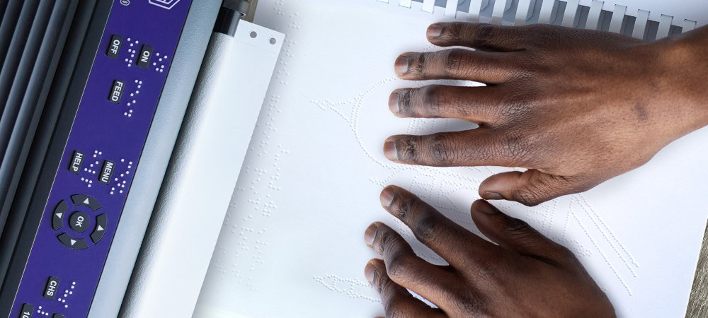 Image of hands resting on a Braille-embossed sheet next to an embosser. The embossed image depicts the Taj Mahal.