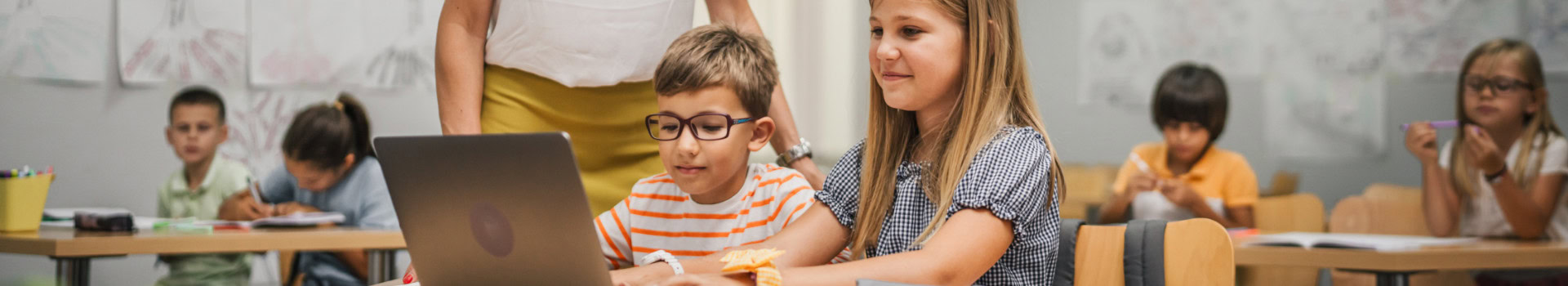 Two children sitting side by side in class, using a laptop, while other students work in the background.