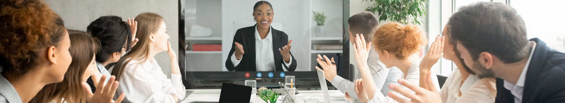Group of colleagues sitting around a table, greeting a woman speaking to them via videoconference on a large screen.