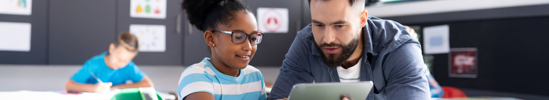 A teacher sitting next to a student, looking together at a digital tablet in class.