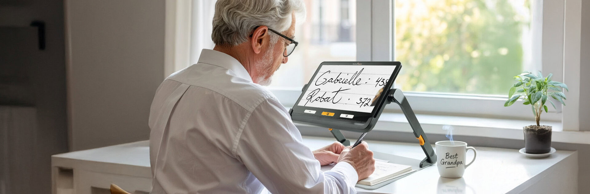 Elderly person using an Explore 12 electronic magnifier to read handwritten text, seated at a desk near a window.