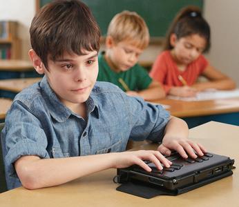 A blind boy uses a braille note taker at his desk in a classroom, while other students write on paper in the background.