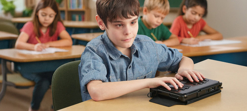 A blind boy uses a braille note taker at his desk in a classroom, while other students write on paper in the background.