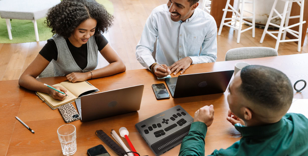 Three people collaborating at a table with laptops and a BrailleNote evolve.