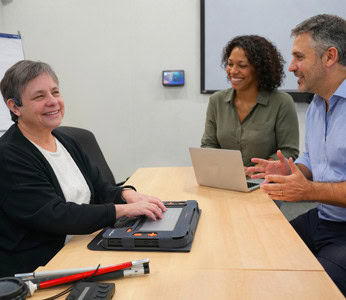 A blind woman uses a braille display during a meeting with two colleagues in a professional office.