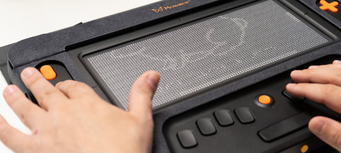 Hands reading braille text on a Monarch tactile display with a small rabbit drawing.