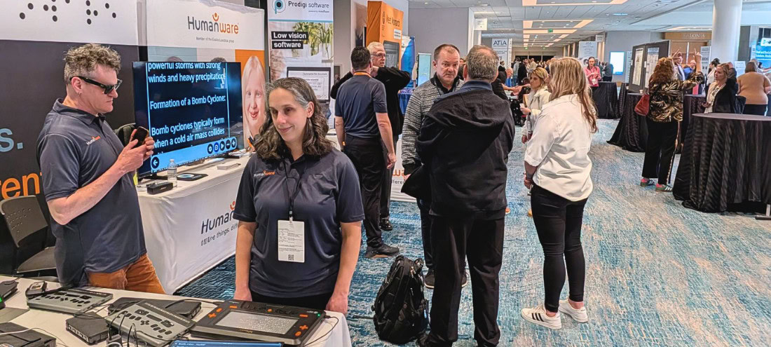 Trade show booth for HumanWare showcasing assistive technologies, with several people talking and demonstrating products in an exhibition hall. Trade show booth for HumanWare showcasing assistive technologies, with several people talking and demonstrating products in an exhibition hall.