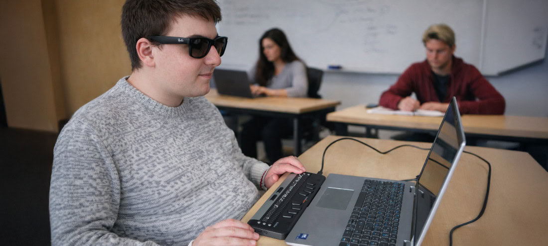 Blind student using a braille display connected to a laptop in a classroom.