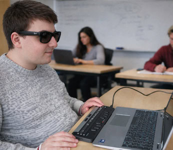Blind student using a braille display connected to a laptop in a classroom.