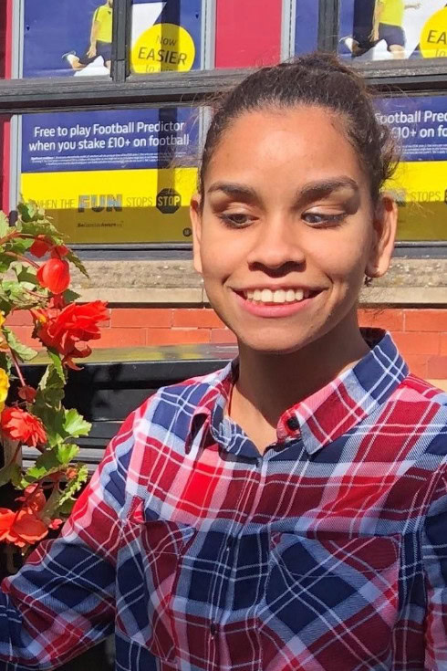 Portrait of Jane smiling outdoors, wearing a red and blue plaid shirt, with red flowers nearby.