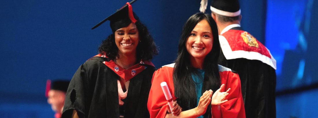 Jane at a graduation ceremony, wearing a black and red gown, smiling on stage beside another graduate.