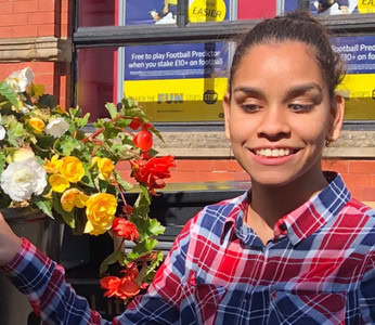 Portrait of Jane smiling outdoors, wearing a red and blue plaid shirt, with red flowers nearby.
