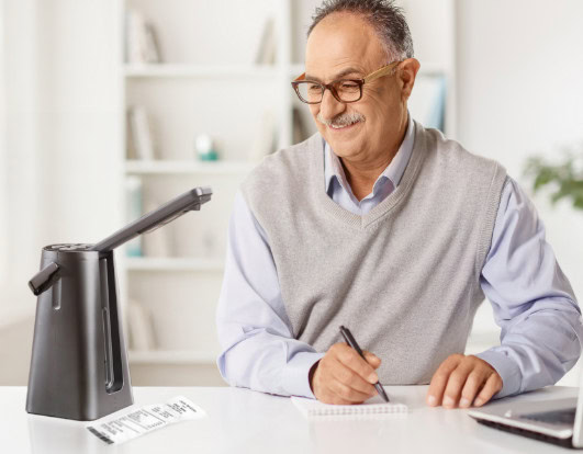 Older man using assistive reading device to review printed receipt and take notes.