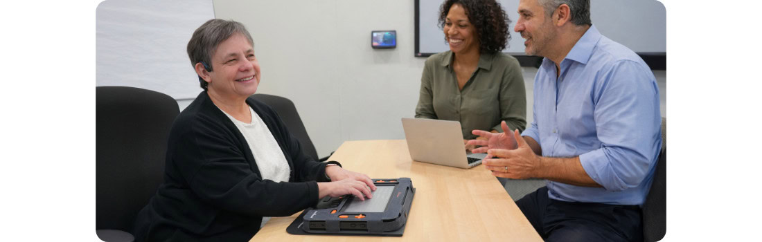 A blind woman uses a braille display during a meeting with two colleagues in a professional office.